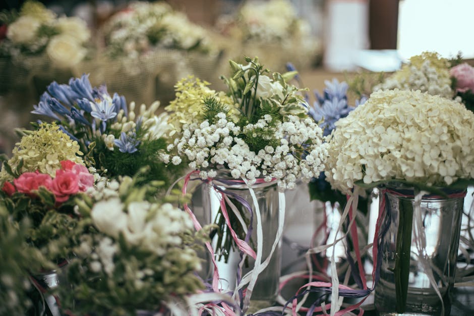 A collection of floral arrangements featuring white hydrangeas, blue anemones, pink roses, and delicate baby's breath, all arranged in tall, clear glass vases wrapped with pink and white ribbons. The 