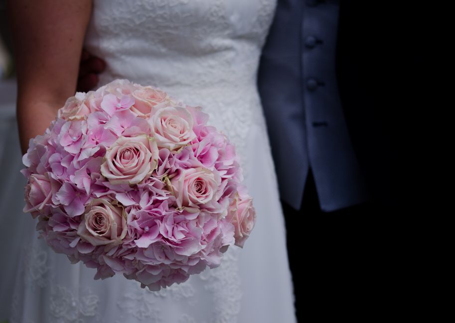 A close-up of a bridal bouquet featuring soft pink roses and pink hydrangeas, arranged in a rounded, lush style. The bouquet is wrapped in light pink ribbon, and held by a bride in a white lace weddin