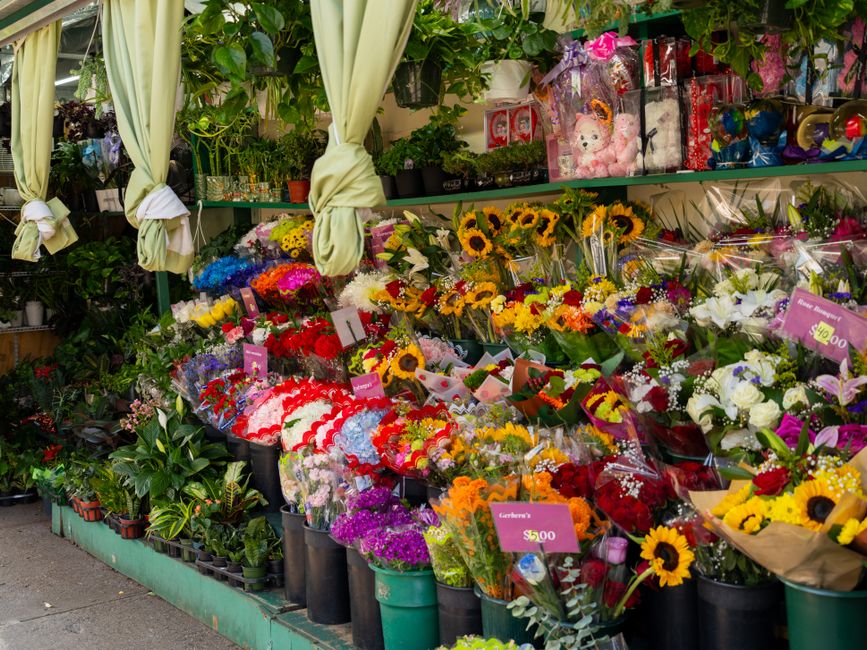 A vibrant flower display at a florist shop featuring an array of fresh floral bouquets arranged on tiered shelves and tables. The bouquets include sunflowers, roses, lilies, carnations, and daisies in