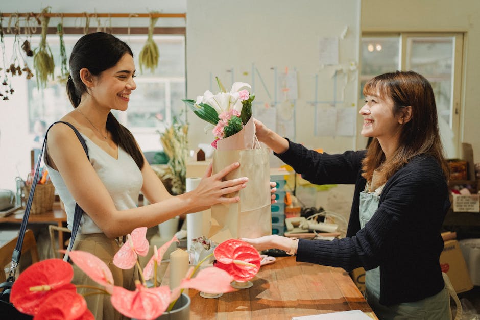 A florist at [COMPANY_NAME] hands a bouquet of fresh flowers, including white and pink blooms, to a smiling female customer inside the shop. The arrangement features a mix of roses and greenery, wrapp